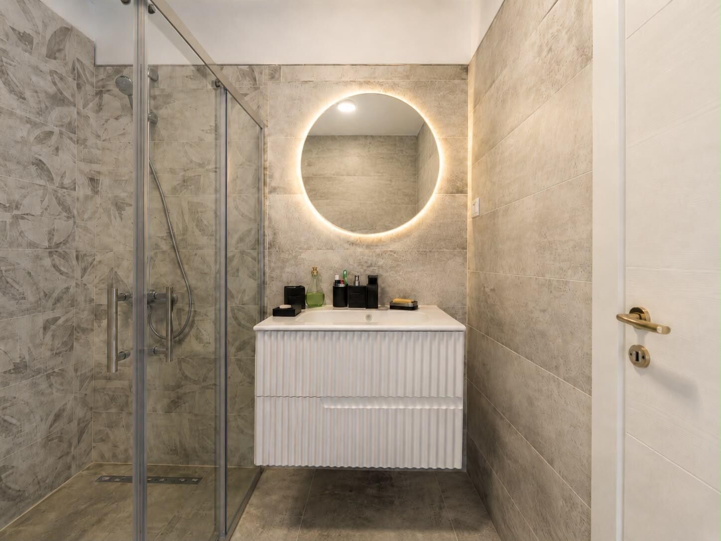 Une salle de bain dotée d’une douche à parois de verre, d’un meuble-lavabo blanc avec un miroir rond et d’une porte avec une poignée en laiton.