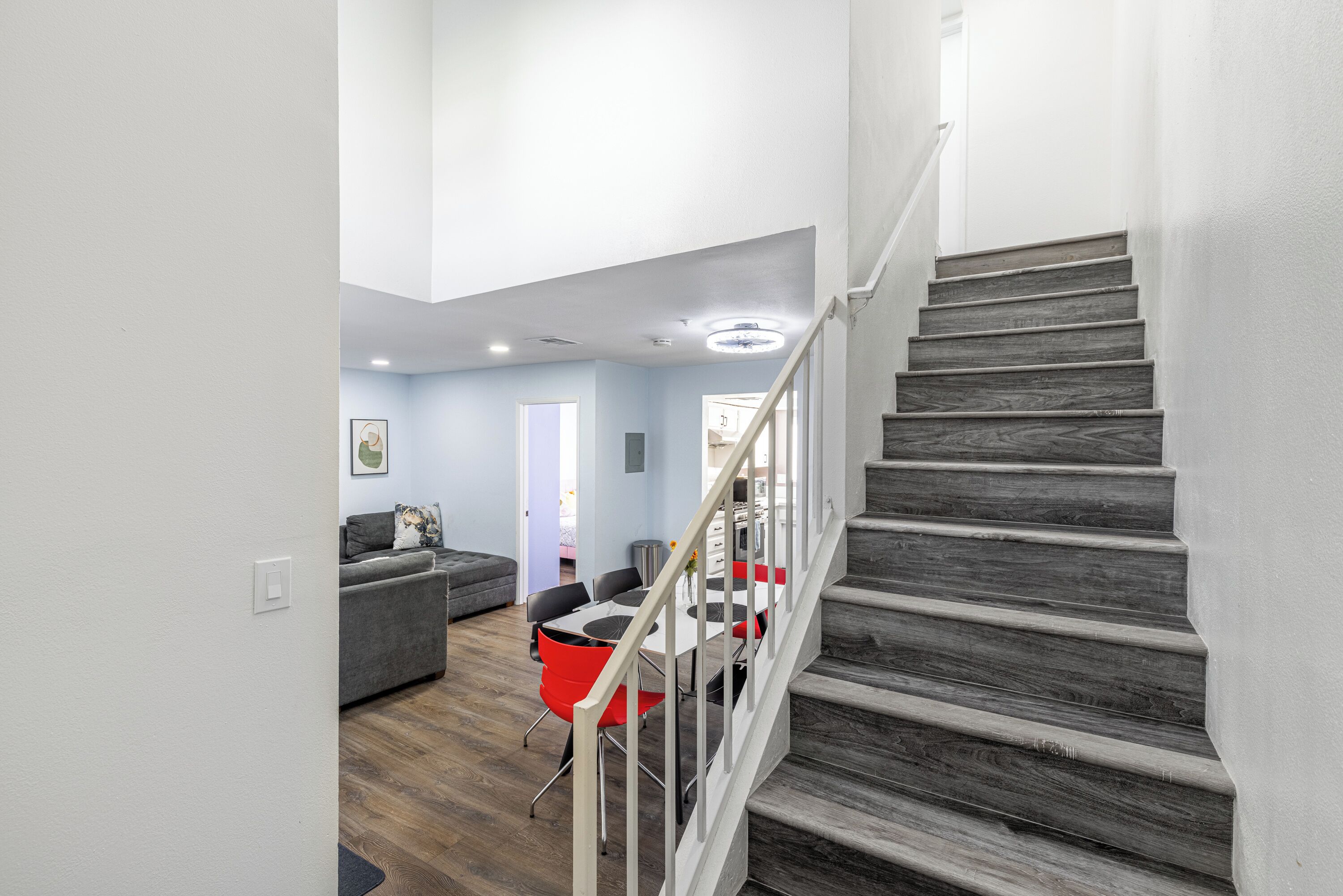 A staircase with wooden steps and a white railing leading to a second floor. A dining area with a red chair and a table is visible in the background.