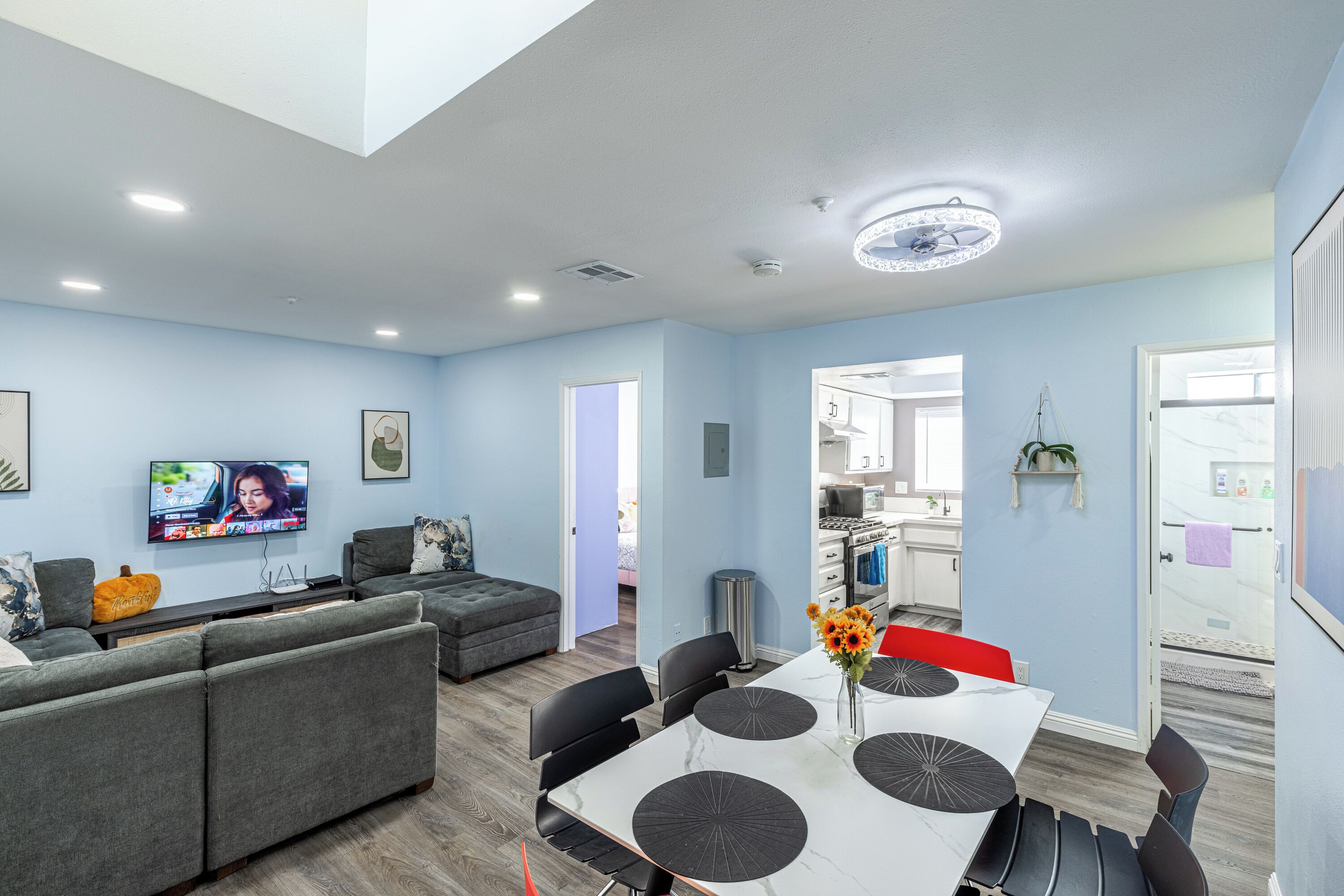 Una sala moderna con un sofá gris, una mesa de comedor con individuales negros y un televisor montado en la pared.