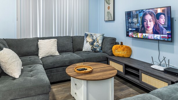 Een moderne woonkamer met een grijze modulaire bank, een rond bijzettafeltje met een mand, een aan de muur bevestigde televisie en een raam met jaloezieën.
