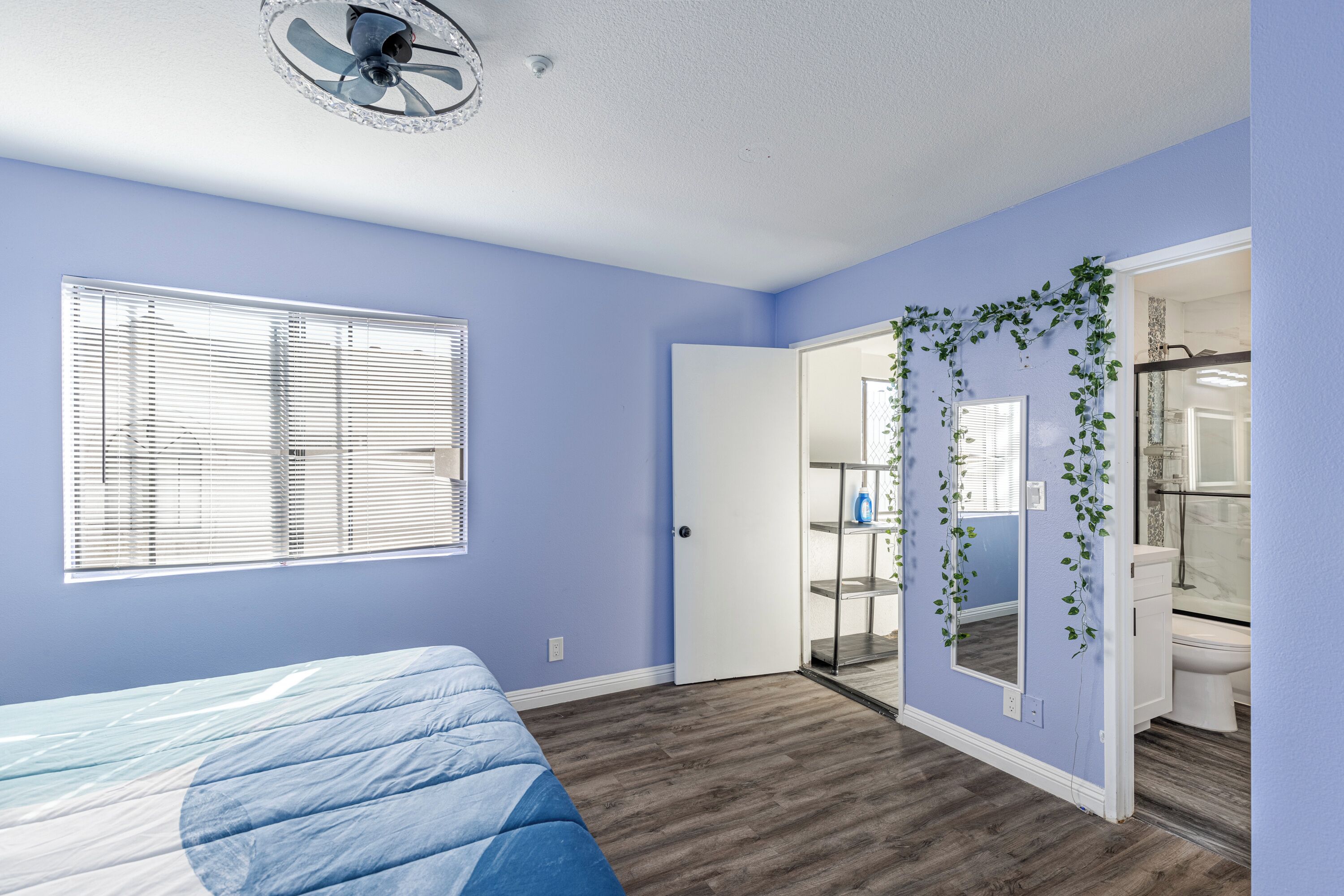 Une chambre à coucher avec un lit, une fenêtre avec des stores, une porte donnant sur une salle de bain et un ventilateur de plafond.