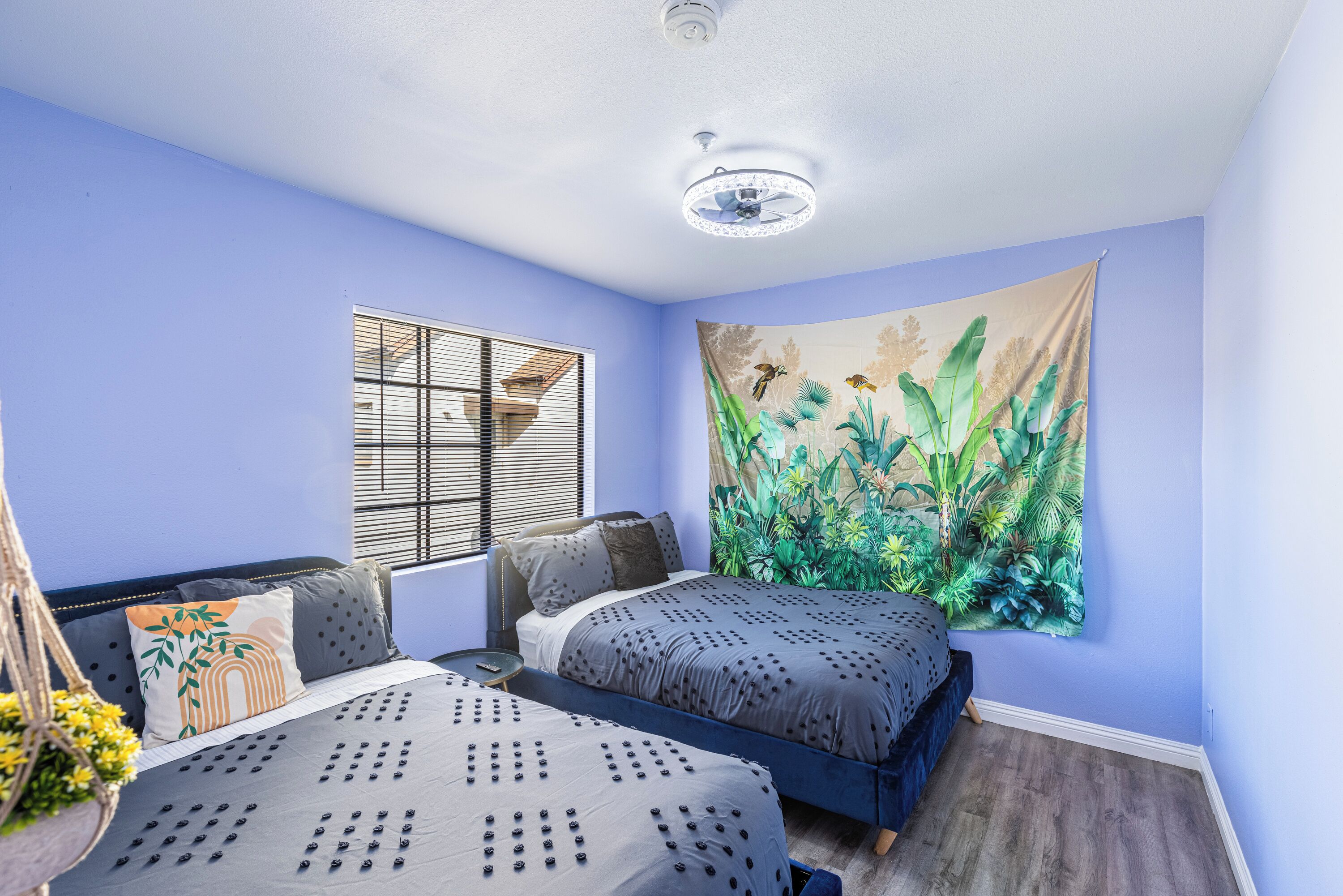 A bedroom with two beds, a window with blinds, a tapestry, and a ceiling fan.