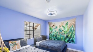 Een slaapkamer met twee bedden, een raam met zonwering, een wandtapijt en een plafondventilator.