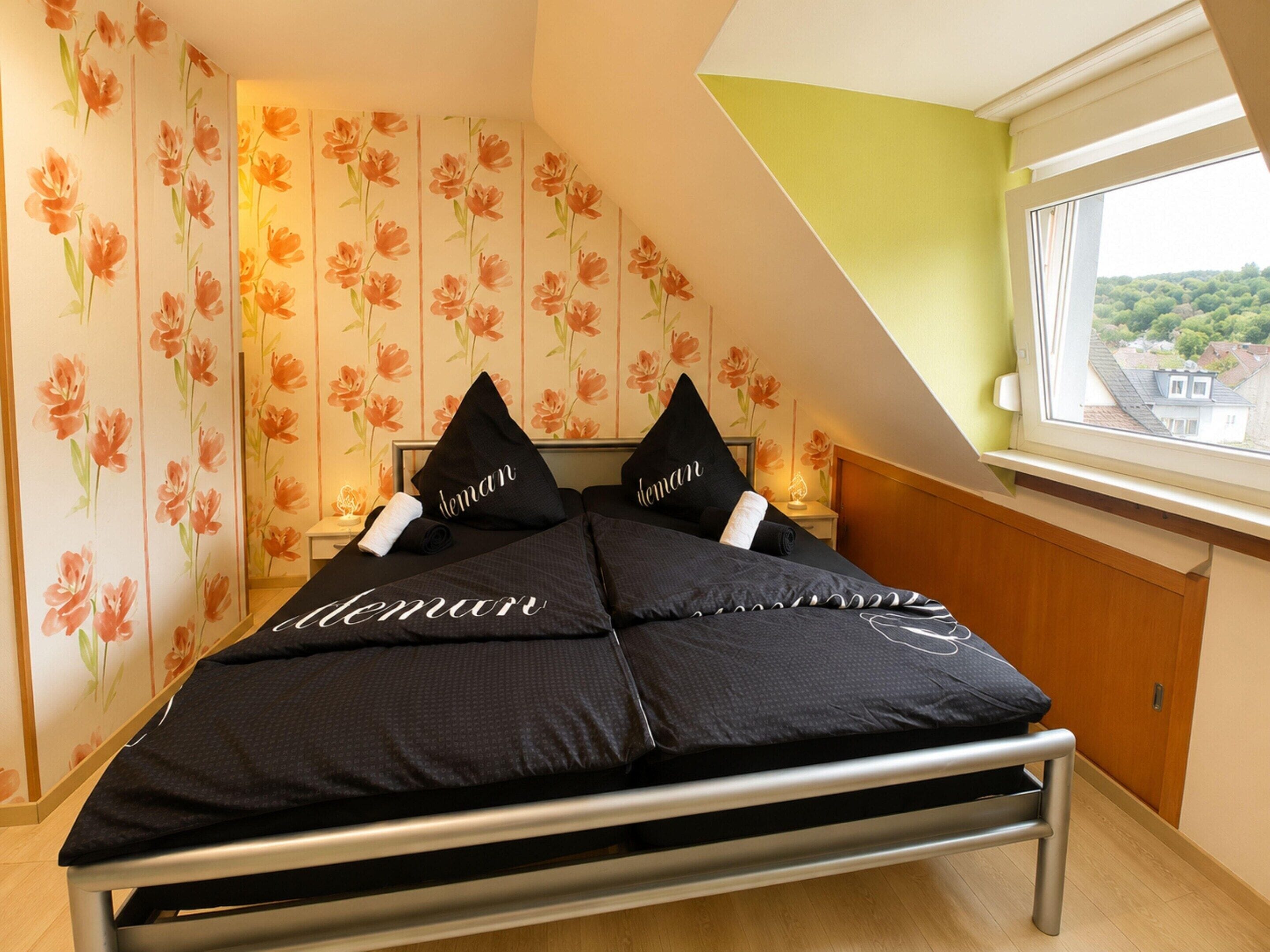 Ein Schlafzimmer mit einem Bett, Kissen, einem Fenster und blumigem Tapetenmuster.