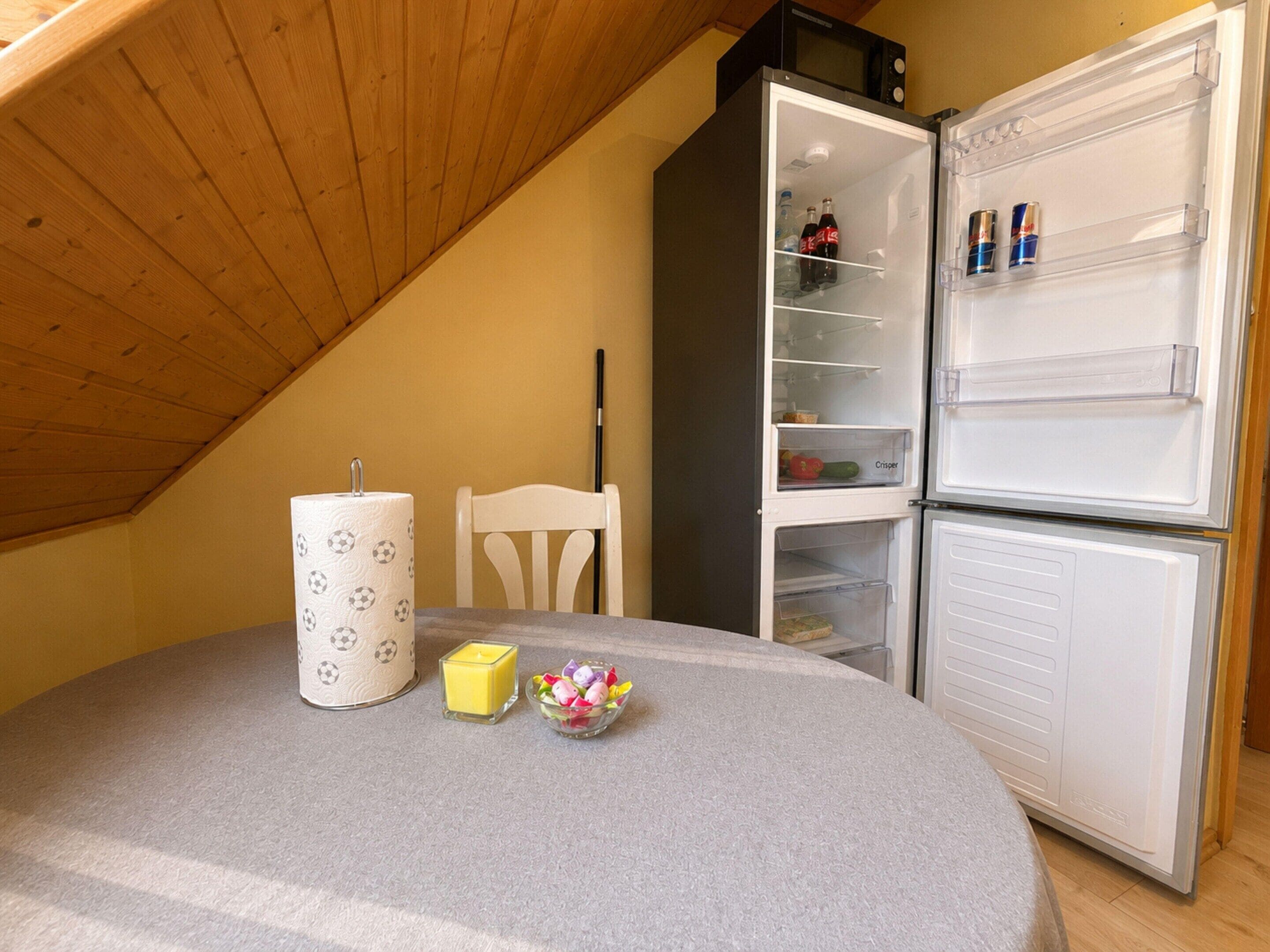 A dining area with a table, a paper towel holder, a candle, and a bowl of fruit. A white chair is positioned next to the table. A refrigerator is visible in the background.