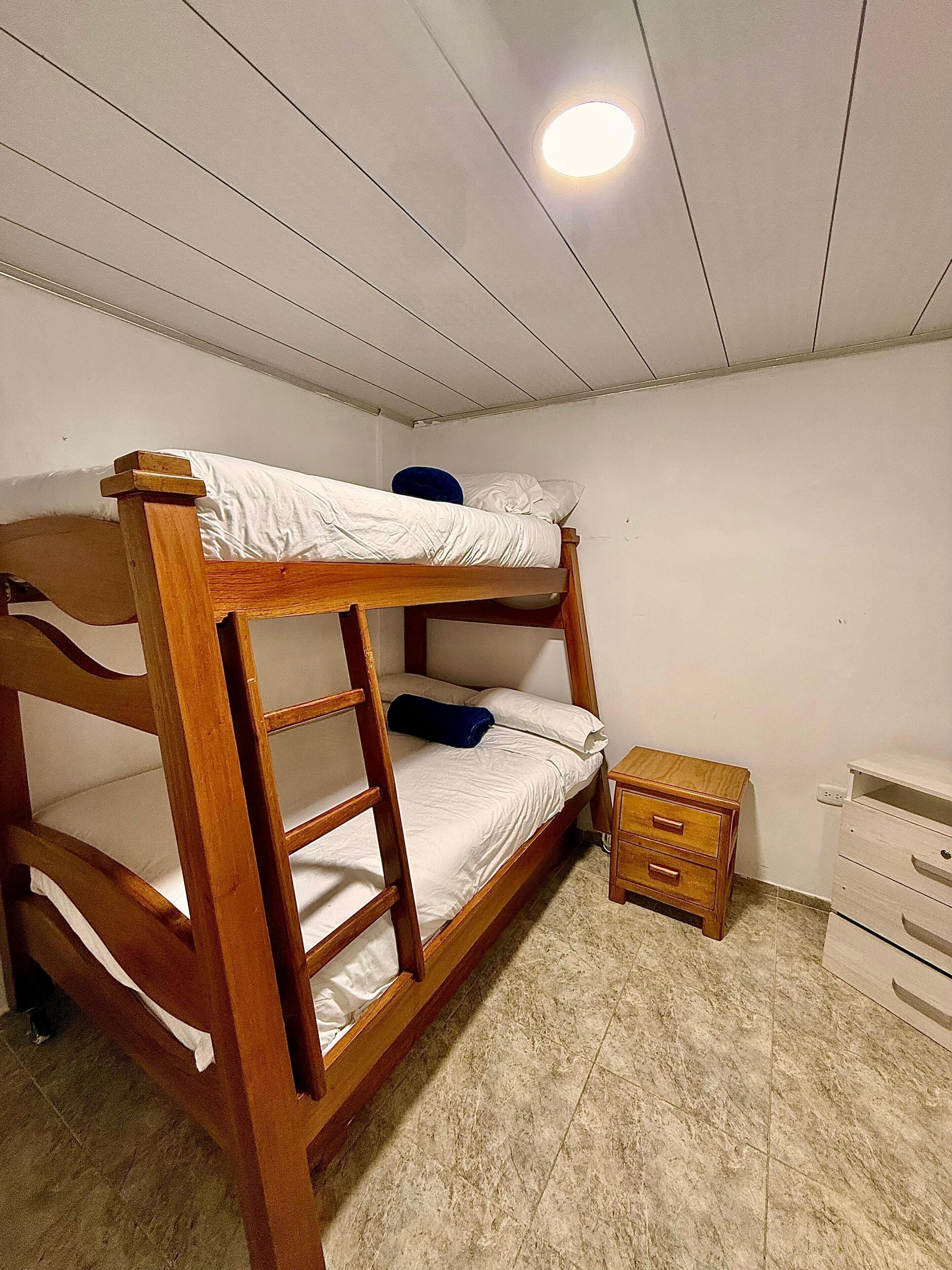 A bunk bed with a wooden frame, a small bedside table, and a dresser in a room with white walls and a tiled floor.