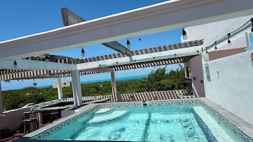 A rooftop swimming pool with a view of the ocean, surrounded by a pergola with hanging lights.