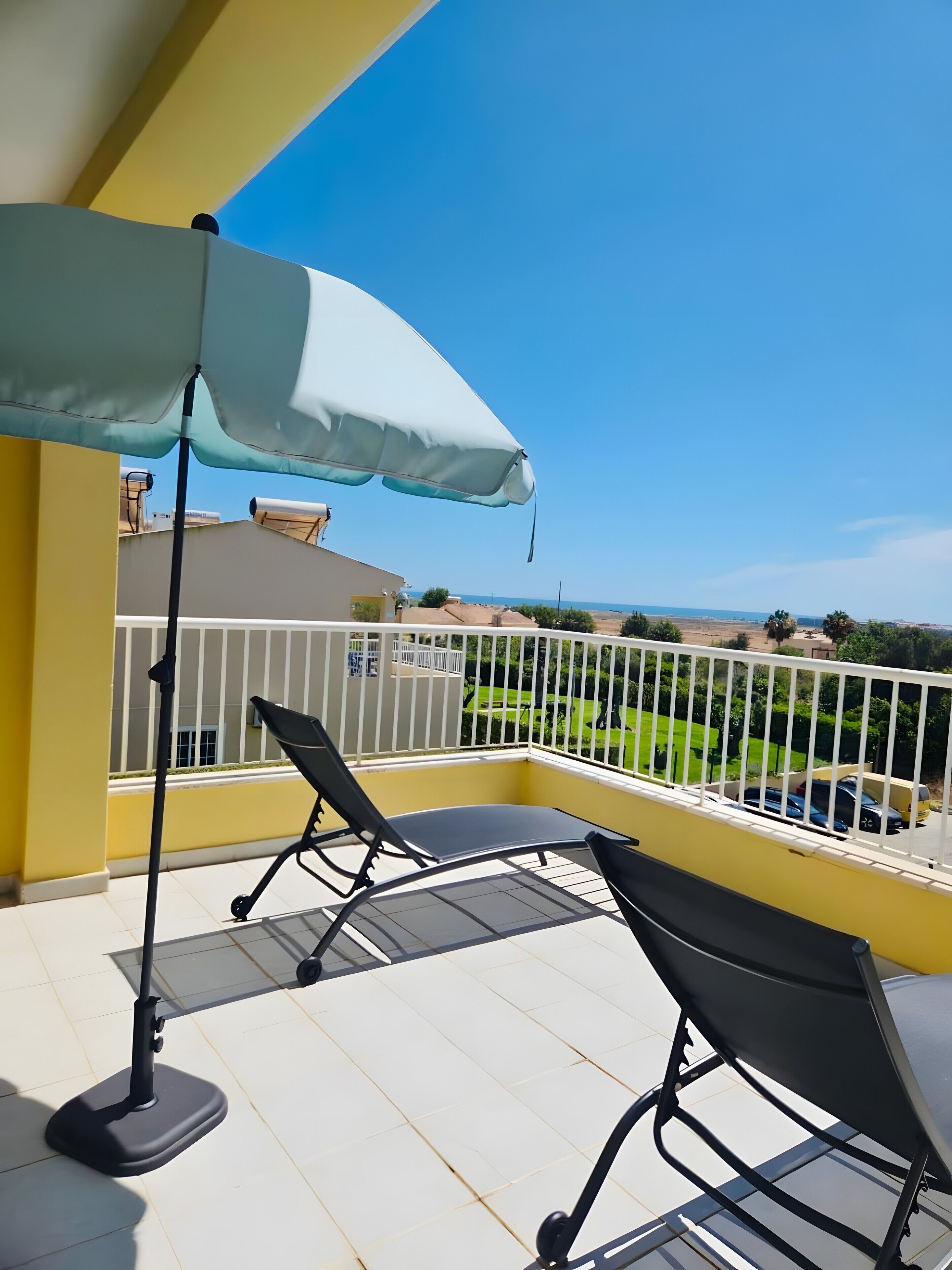Ein Balkon mit Tisch und zwei Stühlen, einem großen Sonnenschirm und Blick auf eine grüne Landschaft sowie entfernte Gebäude.