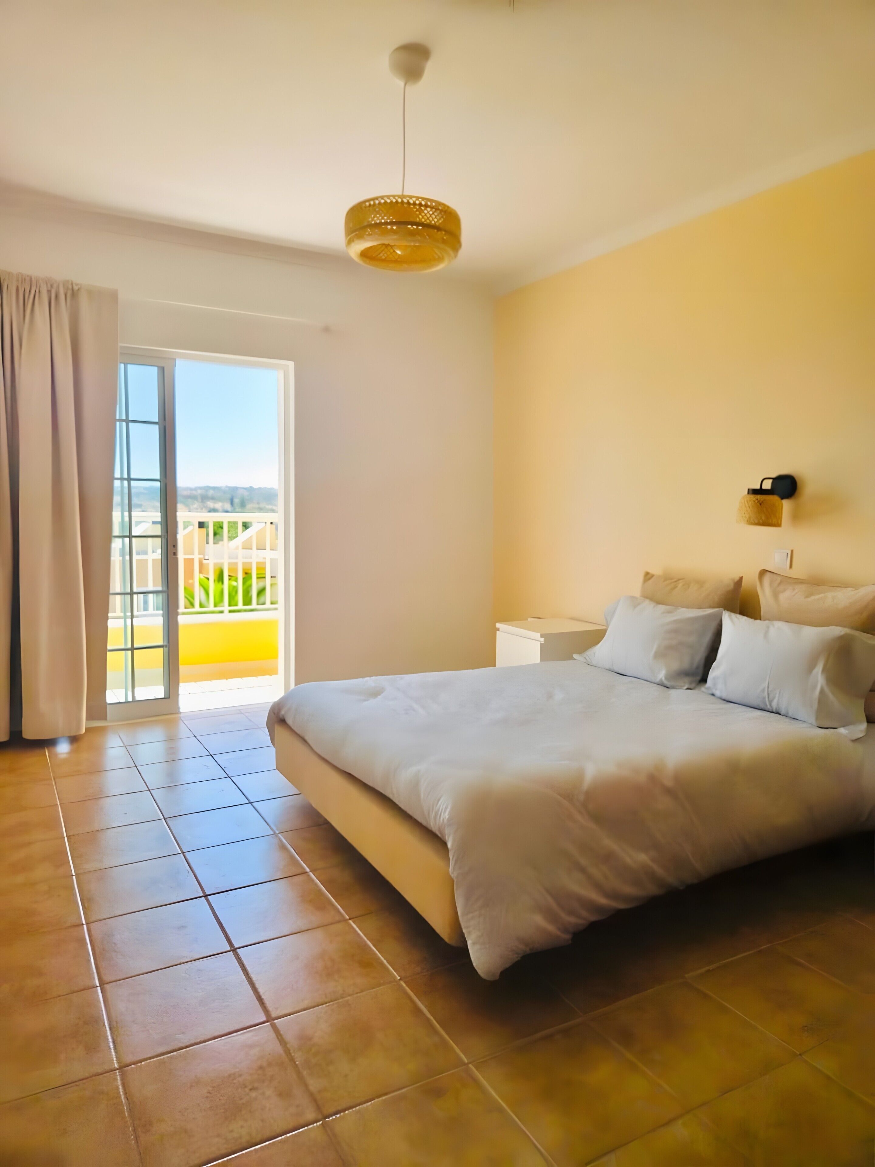 A bedroom with a bed, a nightstand, a wall-mounted lamp, and a view through a sliding glass door.