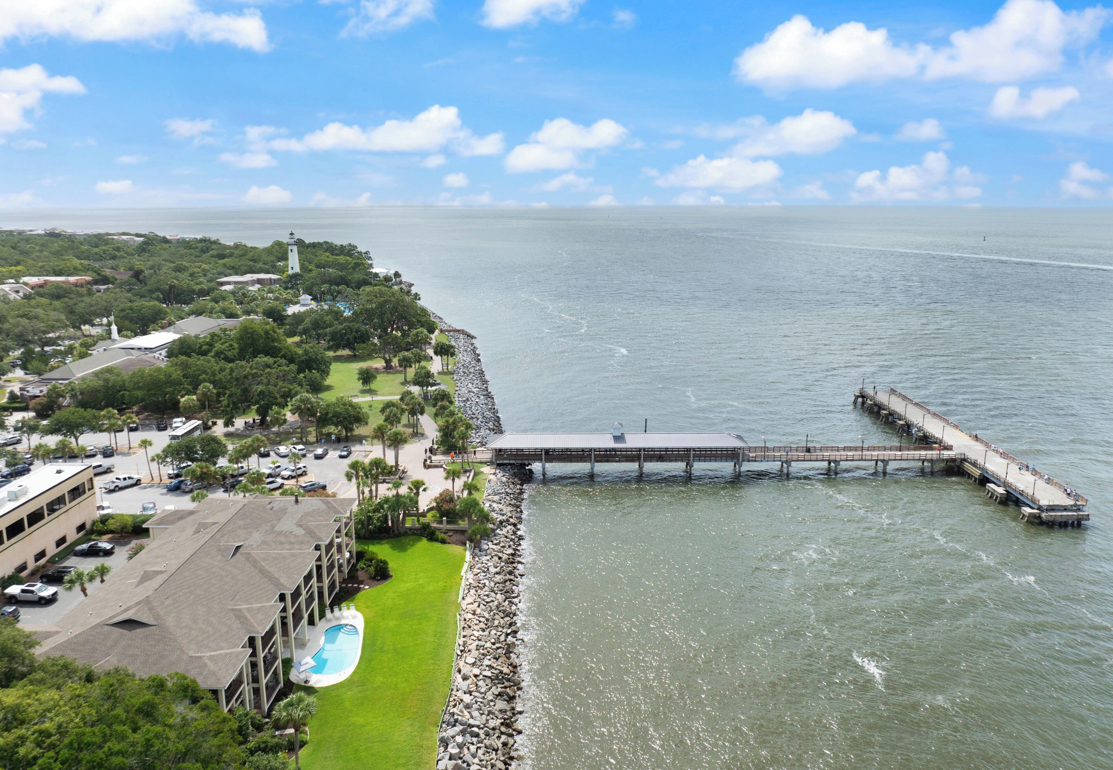 A coastal view with a pier extending into the water, a building with a swimming pool, and a lighthouse in the distance.