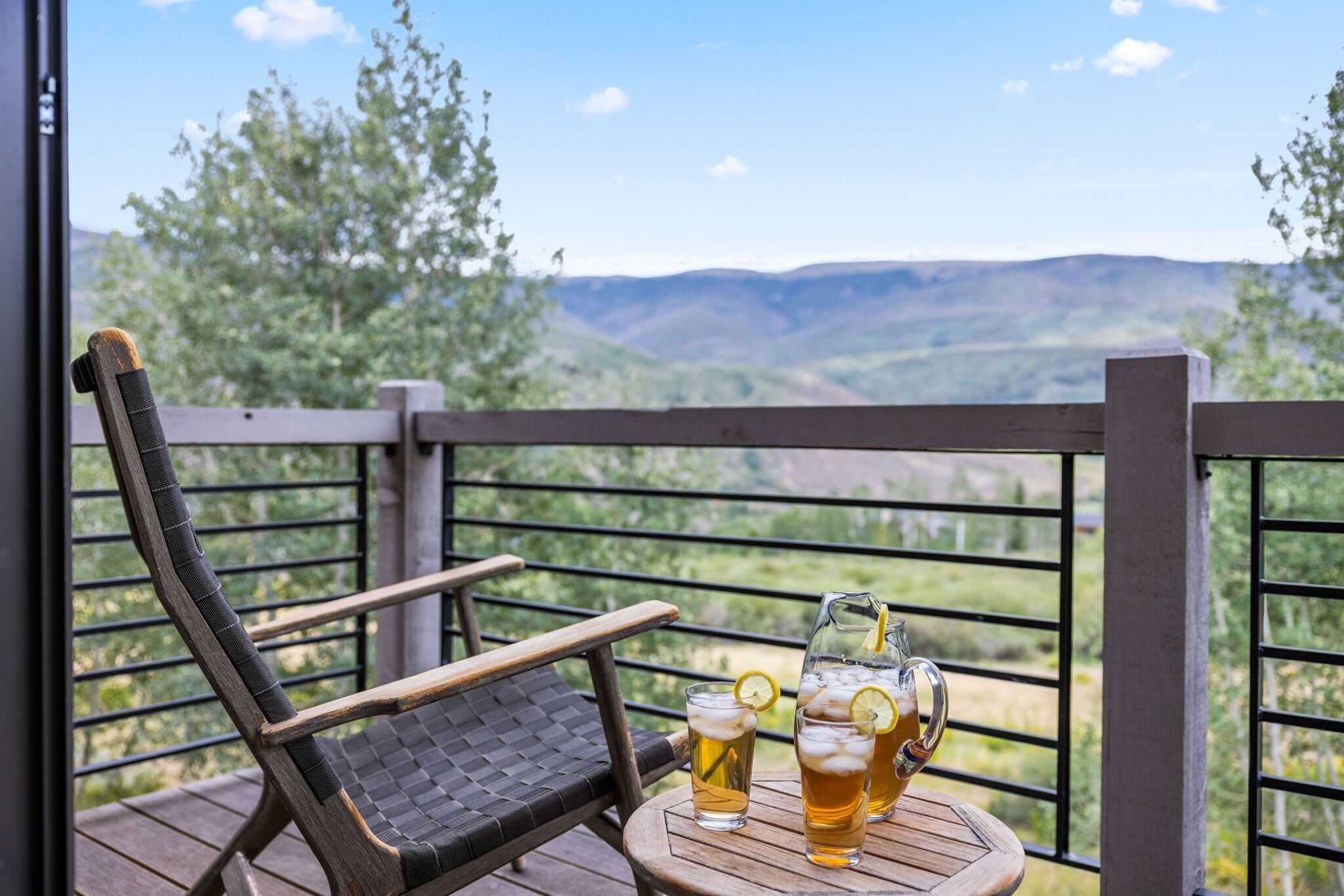 Una silla de madera con baranda metálica, una mesa con dos vasos de cerveza y una jarra, y una vista panorámica de colinas al fondo.