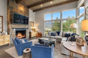 A spacious living room with a stone fireplace, comfortable seating, a flat-screen TV, and large windows offering a view of the outdoors. - NEW! Ultra Luxury Mtn Estate - Hot Tub, Firepits (Silverthorne)