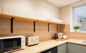 A kitchen with a microwave, toaster, and kettle on the counter, a shelf with cups and glasses above, and a window with a plant. - 125 Columbus Ravine (Scarborough)