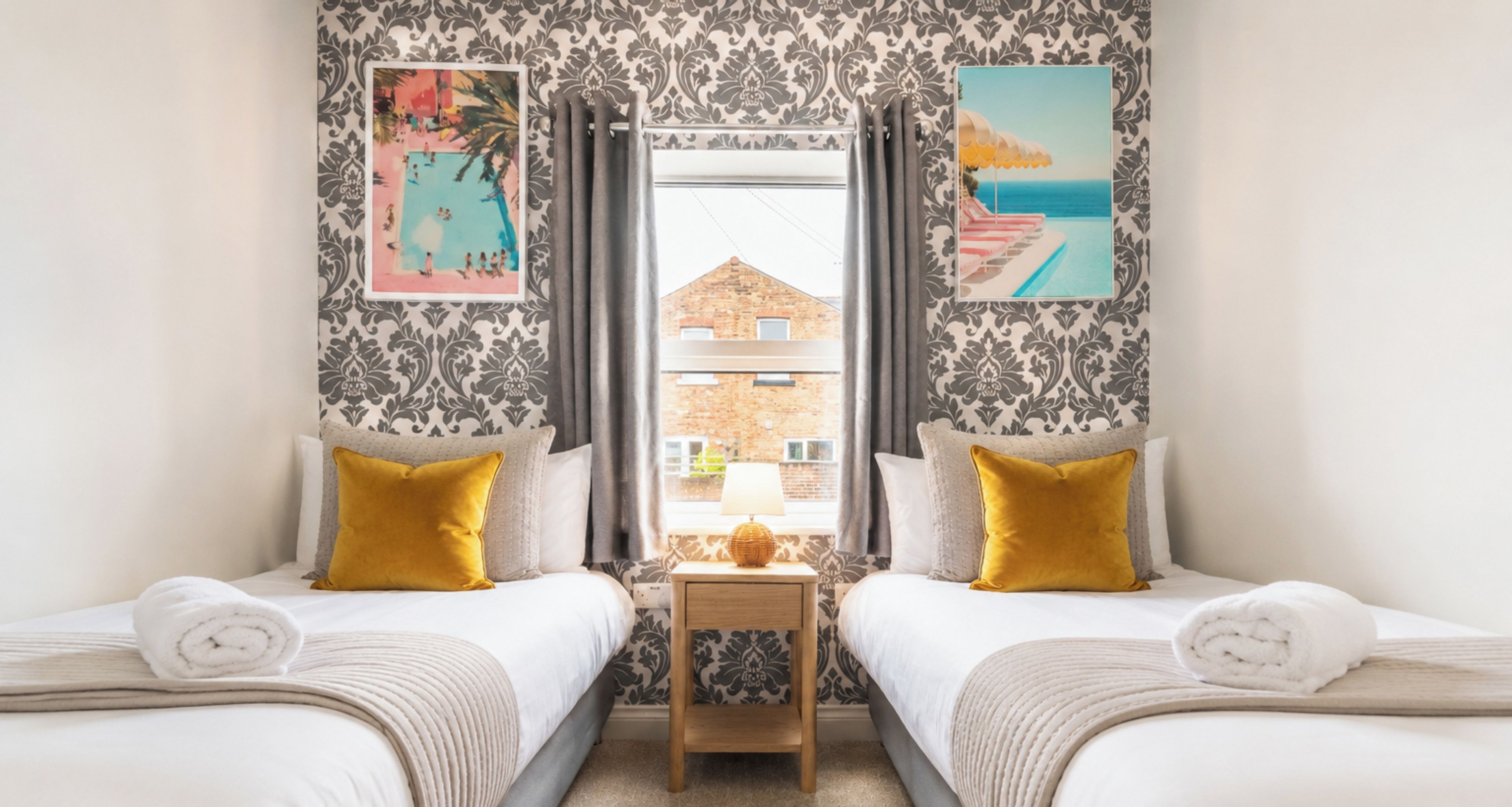 A hotel room with two beds, a small table, and a view of a building through a window.