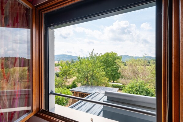 A window with a view of a green landscape, a building with a sloped roof, and a wooden railing.