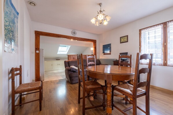 Un comedor con una mesa redonda de madera y sillas, una araña y vista a la cocina.