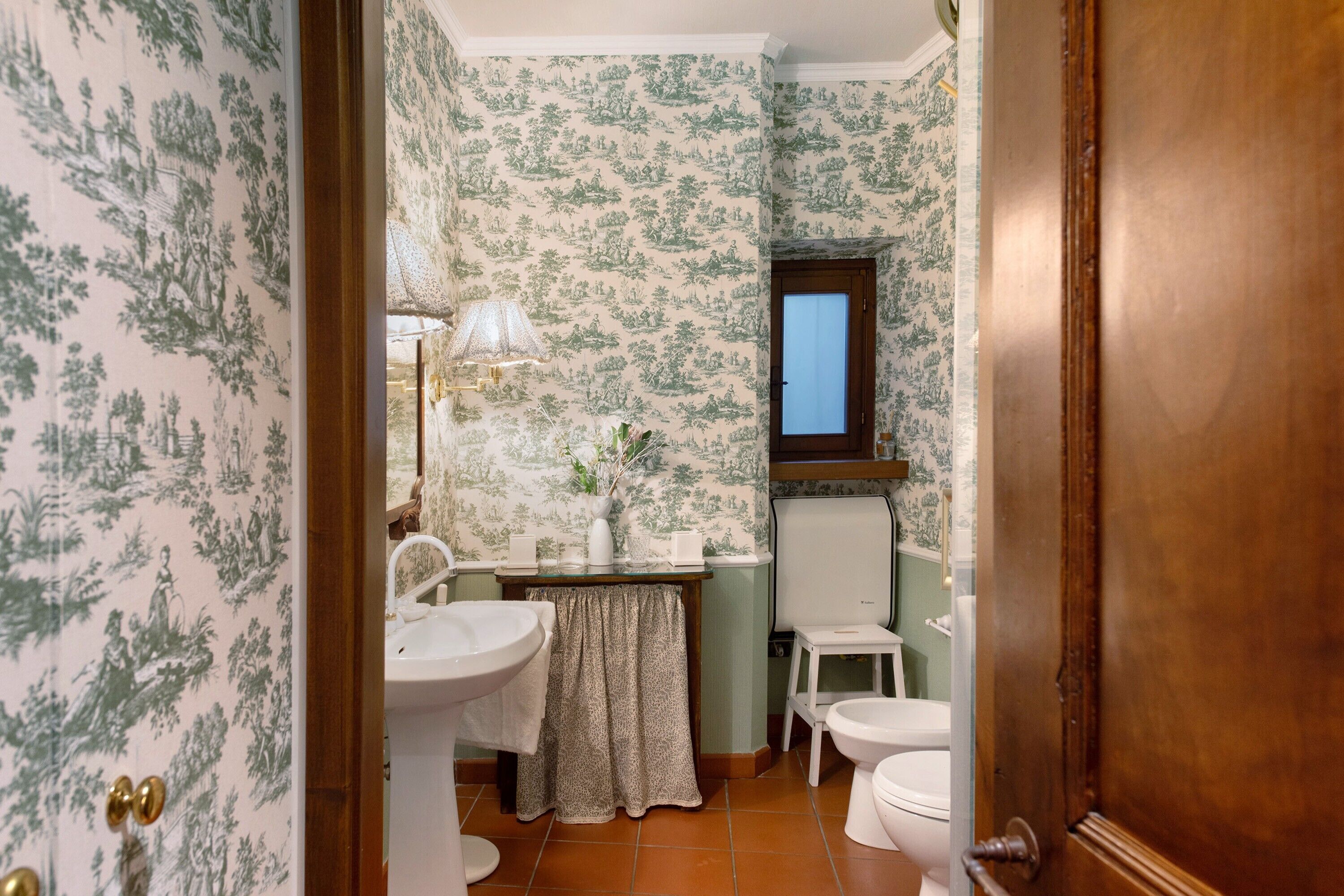 A bathroom with a sink, toilet, and mirror. The walls have a green floral wallpaper.