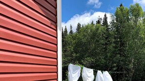A red building with a garden in front, a clothesline with white cloths, and a grassy area with trees in the background. - Rocky Brook Acres (NL)