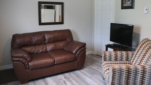 A living room with a brown leather sofa, a patterned armchair, a television on a stand, and a white door.