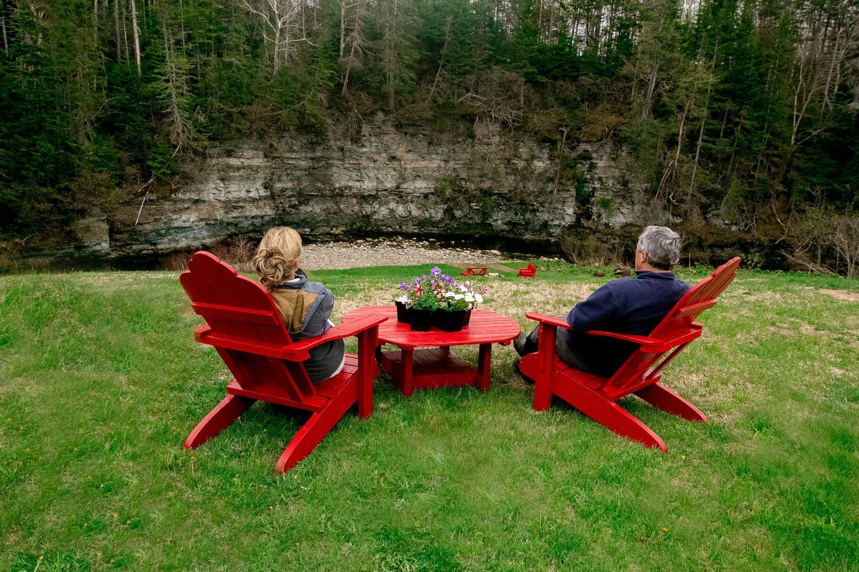 Two people seated at a red table with a flower centerpiece, outdoors with a grassy area and a rocky cliff in the background.