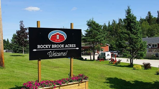 A welcome sign for Rocky Brook Acres with a logo, a wooden planter with flowers, and a clear sky.