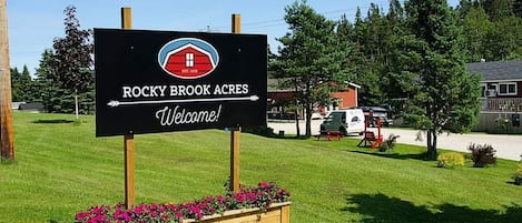 A welcome sign for Rocky Brook Acres with a logo, a wooden planter with flowers, and a clear sky.