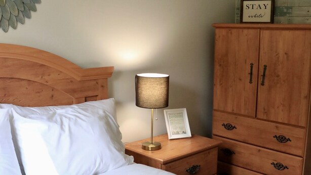A neatly made bed with white linens, a wooden nightstand with a lamp and a framed certificate, and a wooden cabinet with a 'STAY' sign above it. - Rocky Brook Acres (NL)