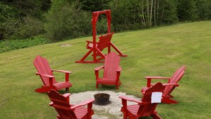 A set of red Adirondack chairs arranged around a fire pit in a grassy outdoor area. - Rocky Brook Acres (NL)