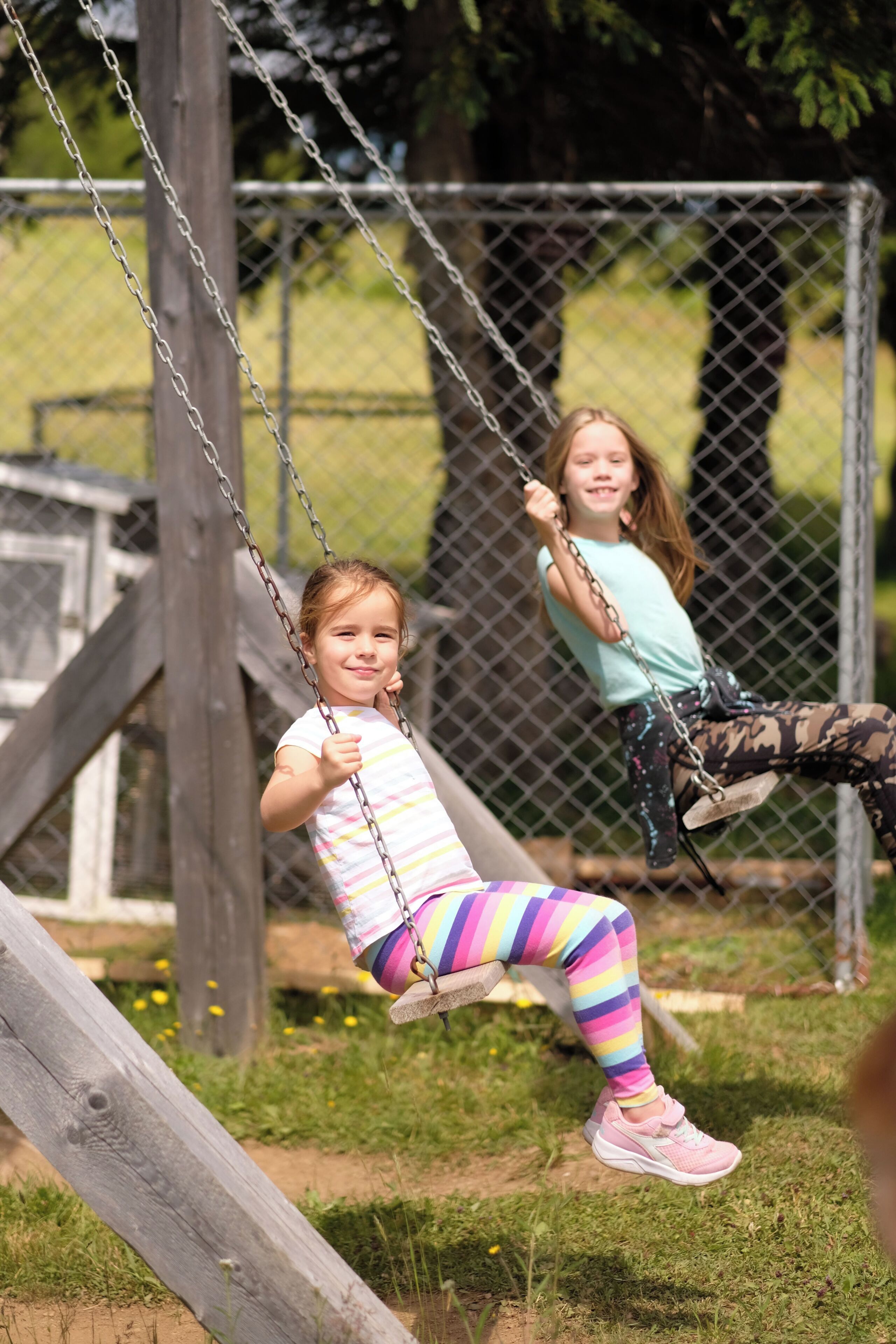 Two children on swings at a playground with a wooden structure and chain-link fence in the background.