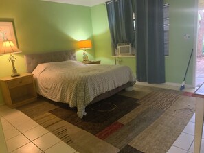 Ein Schlafzimmer mit Bett, Nachttisch, Lampe, Klimaanlage und einem Fenster mit Vorhängen.