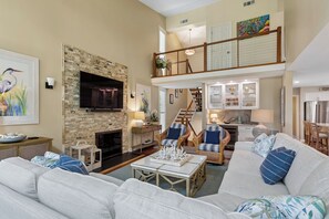A spacious living room with a fireplace, a television, and a dining area with a kitchen in the background.