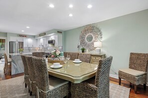 A dining area with a glass-top table surrounded by woven chairs, a decorative mirror, and a view of the kitchen in the background.