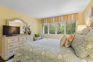 A bedroom with a bed, dresser, television, window with curtains, and a view of greenery outside.