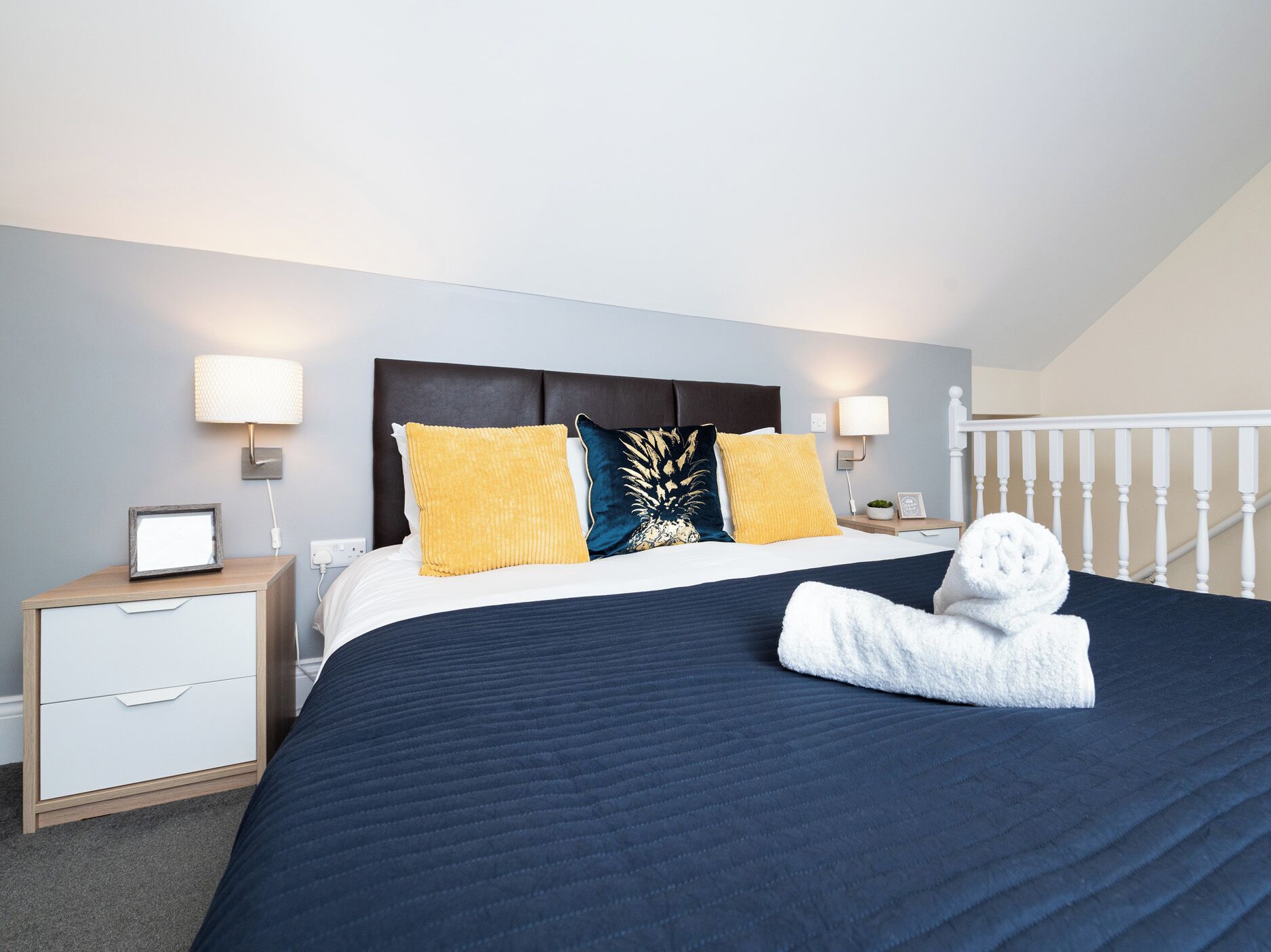 A hotel room with a bed, two bedside tables, a nightstand, a lamp, and a white railing.