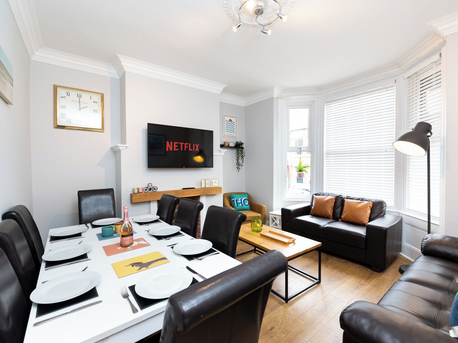 A modern living room with a dining area, a sofa, a TV mounted on the wall, and a window with blinds.