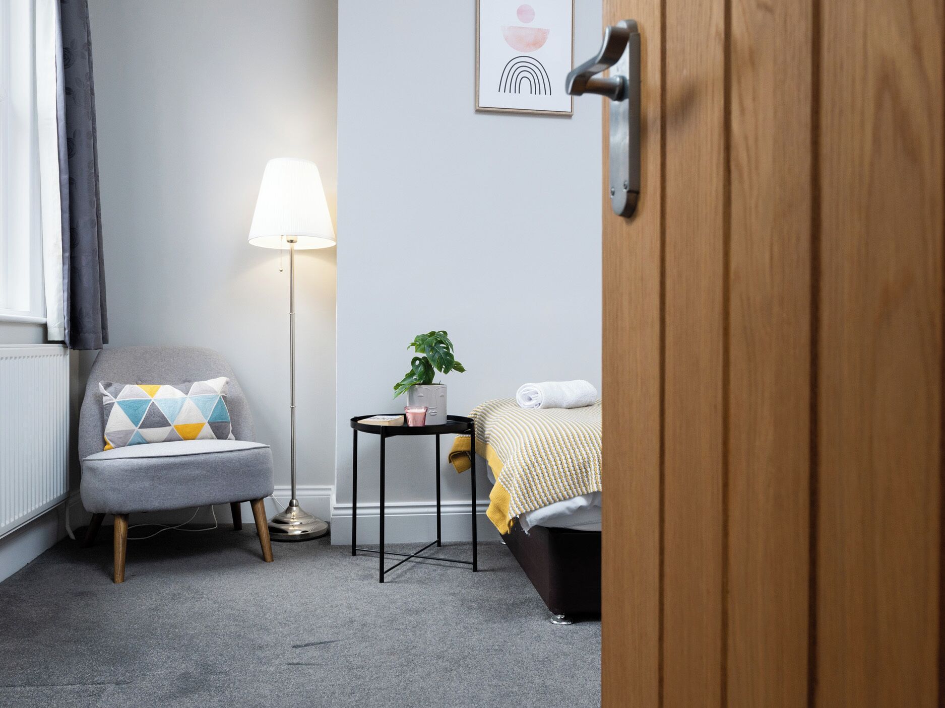 A hotel room with a grey armchair, a floor lamp, a small table with a plant, and a bed with a yellow blanket.
