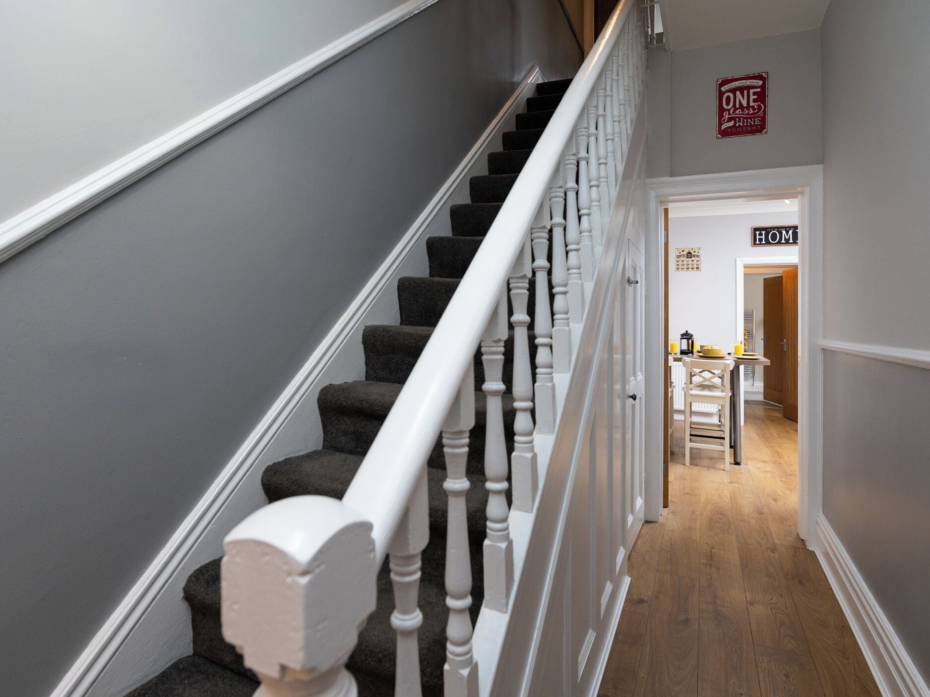 Una escalera con balaustres blancos y peldaños alfombrados que conducen a una habitación con piso de madera y una mesa con un letrero que dice 'HOME'.