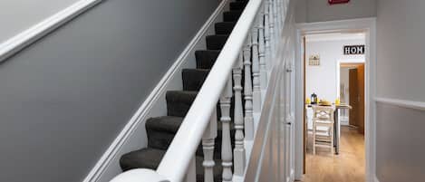 Una escalera con balaustres blancos y peldaños alfombrados que conducen a una habitación con piso de madera y una mesa con un letrero que dice 'HOME'.