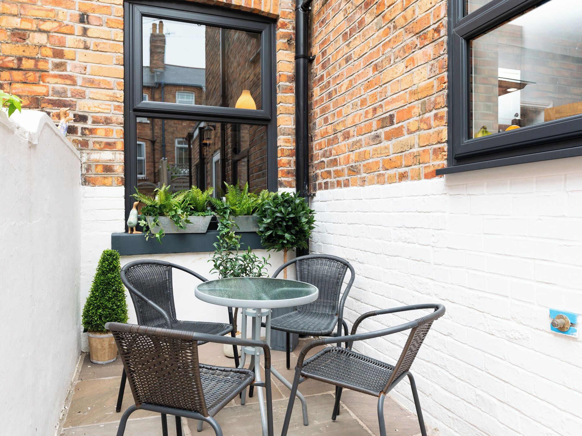 A small outdoor seating area with a round table and four chairs, surrounded by potted plants and a white brick wall.