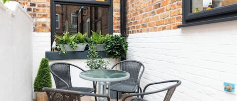 A small outdoor seating area with a round table and four chairs, surrounded by potted plants and a white brick wall.