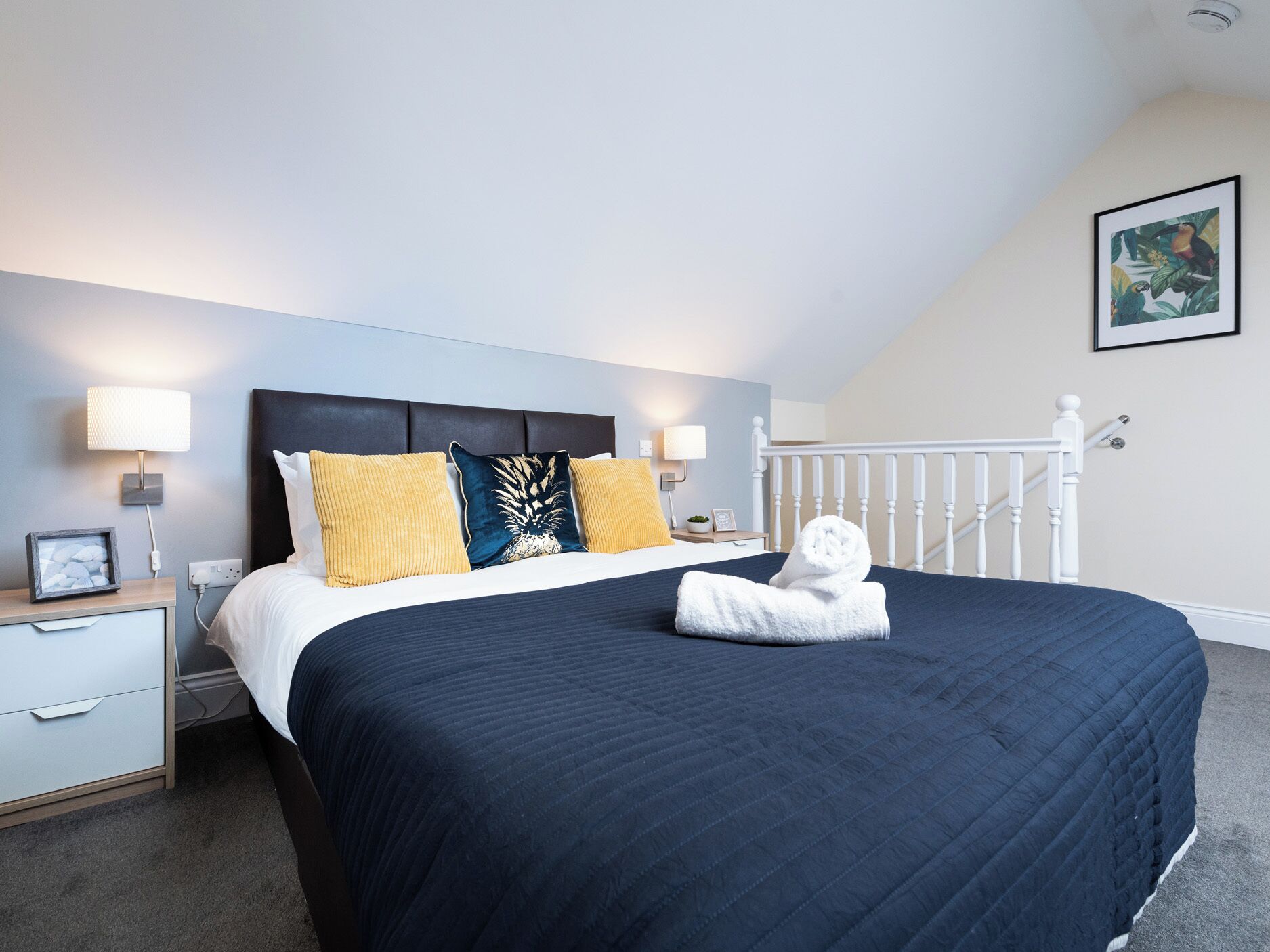 A hotel room with a bed, bedside lamps, a nightstand, a framed picture, and a white railing.