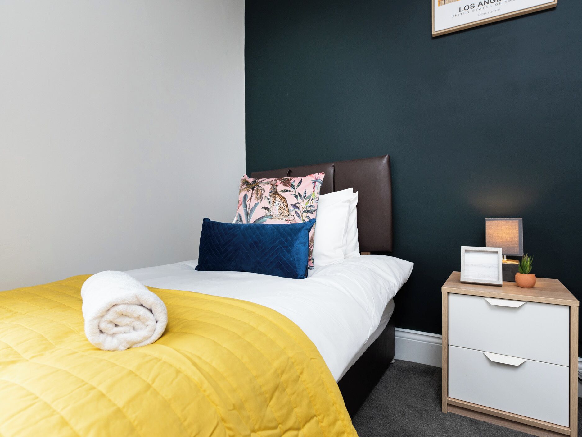 A neatly made bed with a yellow bedspread, a white pillow, and a blue pillow with a floral design. There is a white nightstand with a lamp and a small plant beside the bed.