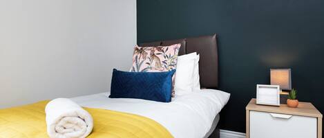 Una cama bien hecha con una colcha amarilla, un almohadón blanco y un almohadón azul con diseño floral. Junto a la cama hay una mesita de noche blanca con una lámpara y una pequeña planta.