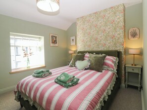 A bedroom with a bed, two bedside tables, a window with blinds, and a floral-patterned wall. - Foxglove Cottage (Conwy)