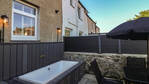 A spa area with a hot tub, a table, and chairs, adjacent to a building with a window and a stone wall. - Foxglove Cottage (Conwy)