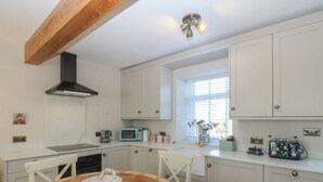 A kitchen with white cabinets, a wooden ceiling, a round table with chairs, and a window with blinds. - Foxglove Cottage (Conwy)