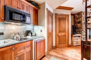 A kitchen with wooden cabinets, a microwave, a kettle, and a stone accent wall. - BC Slopeside Ski Studio, Hot Tub, Resort Amenities (Beaver Creek)