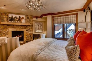 A bedroom with a stone fireplace, a bed with patterned pillows, a chandelier, and a window with a view. - BC Slopeside Ski Studio, Hot Tub, Resort Amenities (Beaver Creek)