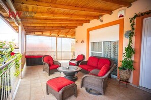 A covered outdoor seating area with wicker furniture and red cushions, a round table, and a potted plant. - Escape to our spacious country home. (DIMOS EUROTA)