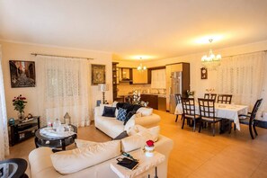 A living room with a sofa, a dining table with chairs, a kitchen area, and a chandelier. - Escape to our spacious country home. (DIMOS EUROTA)